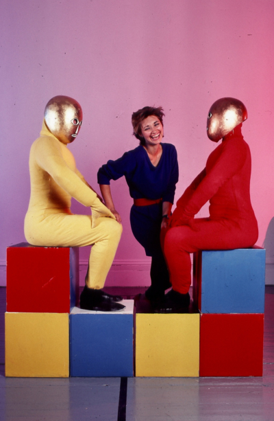 Baukastenspiel rehearsal. Two dancers are sitting in profile, facing each other. The dancer on the left wears a yellow jumpsuit, and the one on the right wears a red one. The dancers are wearing golden masks that cover their entire heads. They are sitting on cubes stacked two rows high. Debra McCall is in the background smiling.