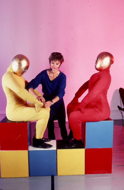 Baukastenspiel rehearsal. Two dancers are sitting in profile, facing each other. The dancer on the left is wearing a yellow jumpsuit, and the one on the right is a red jumpsuit. The dancers are wearing golden masks that cover their entire heads. They are sitting on cubes stacked two rows high. Debra McCall is in the background, adjusting the dancer's costume on the left.