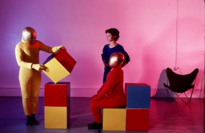 Baukastenspiel rehearsal. One dancer in a yellow jumpsuit standing on the left is holding a cube stacked on two other cubes. To the right, another dancer wearing a red jumpsuit is in profile, sitting on a cube. The dancers are wearing golden masks that cover their entire heads. The faces of the cubes are solid colors: red, yellow, or blue. Debra McCall is standing in the background.