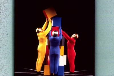 Baukastenspiel performance. Three performers in solid yellow, red, or blue jumpsuits reach and stack cubes high on the stage floor. All of them are wearing golden masks that cover their entire heads.