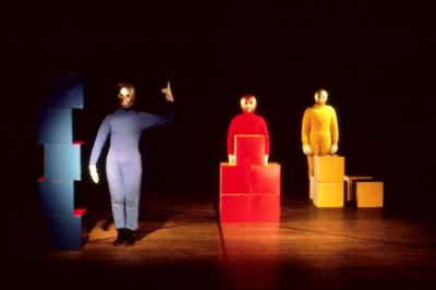 Baukastenspiel performance. Three performers stand on stage, each wearing a blue, red, or yellow jumpsuit from left to right. They all wear golden masks that cover their entire heads. The performer on the left, in blue, is at the front of the stage, standing next to a stack of blue cubes, pointing up with his index finger. In red, the performer in the middle is at the center of the stage, standing behind a stack of red cubes that reach up to his chest. The performer on the right, in yellow, is at the back of the stage, standing behind yellow cubes that reach up to his hips.