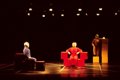 Baukastenspiel performance. Three performers are on stage. They are dressed in a blue, red, or yellow jumpsuit from left to right. All of them are wearing golden masks that cover their entire heads. In blue, the performer on the left sits in profile on stacked red cubes, facing the right of the image. The performer in the middle faces the viewer, dressed in red, and sits on red cubes staked to form an armchair. The performer on the right is wearing a yellow jumpsuit, standing with one foot on a yellow cube, leaning on a stack of yellow cubes, and resting his hand on the stack.