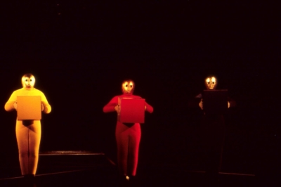 Baukastenspiel performance. Three performers stand in line on stage, facing the viewer. All of them are wearing golden masks that cover their entire heads. The one on the left is wearing a yellow jumpsuit and holding a yellow cube up to his chest. The performer in the middle is doing the same, dressed in a red jumpsuit and holding a red cube. The last performer is in blue, holding a blue cube.