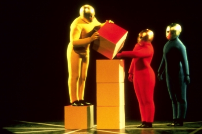 Baukastenspiel performance. Three performers are standing on stage, dressed in solid yellow, red, or blue jumpsuits. All of them are wearing golden masks that cover their entire head. On the left, a performer stands on a yellow cube and places a fourth cube on a three-cube high stack—the performers on the right help.