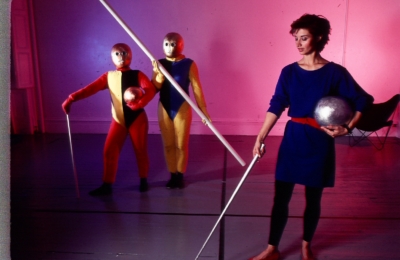 Formentanz rehearsal. Two dancers are standing on the left in the background, and Debra McCall is on the right. All three subjects are holding staffs. The dancers are wearing multicolored jumpsuits in red, yellow, and blue, with golden masks that cover their entire heads.