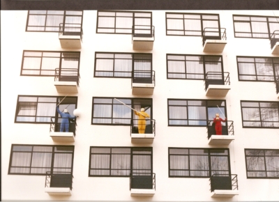 Image of a white apartment building showing rows of apartment floors. Three balconies are on the third row of apartments, each with one performer. Each performer wears a blue, yellow, and red jumpsuit from left to right. All performers also wear golden masks that cover their entire heads. The first performer on the left holds up a wand with his right hand and a white sphere with his left hand. The performer in the middle holds a pole across his body with both hands, and the performer on the right has a white wand and a small metallic sphere.