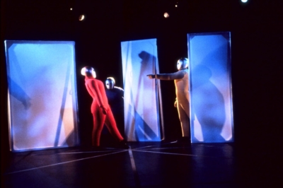Baukastenspiel performance. Three performers are on stage wearing red, blue, and yellow jumpsuits. Each of them has a golden mask covering their entire head. The space is divided by three portable partitions. The performer on the right side, wearing yellow, stands between two of these partitions and points to the performer on the left, who is wearing a red jumpsuit and leaning back. The performer in the middle, wearing blue, is in the background and looking at the red performer.
