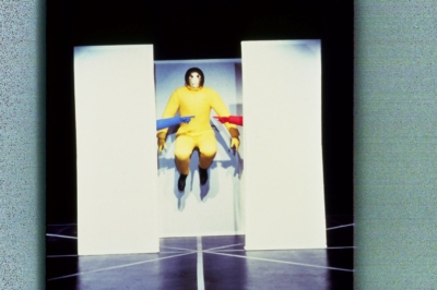 Baukastenspiel performance. Three white partitions are in the middle of the image. A performer in a yellow jumpsuit jumps in the image's center. Two hands, one red and one blue, with pointing index fingers, appear behind the left and right partitions.