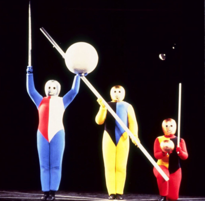 Gestentanz performance. Three performers are wearing multicolored jumpsuits. Two of them are standing on the left while the one on the right kneels. All of them are wearing golden masks that cover their entire head. The performer on the left holds a wand with his right hand and a white sphere with his left hand. The one in the middle is holding a pole diagonally across the front of his body. The dancer on the left has a small golden sphere in his right hand and a wand in his left hand.