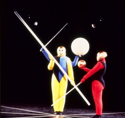 Gestentanz performance. Three performers wearing multicolored jumpsuits. Two performers, one in front of the other, face the viewer, while the one on the right stands in profile, facing the left of the image. All of them are wearing golden masks that cover their entire head. The performer in the foreground holds a pole diagonally across the front of his body. The performer behind him has a sphere and a wand; only his hands are visible. The performer on the right holds a small golden sphere and a wand in his other hand, pointing down.