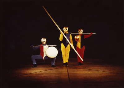 Gestentanz performance. Three performers are in line in front of the viewer. They are wearing multicolored jumpsuits. All of them are wearing golden masks that cover their entire head. The one on the left is squatting and holds a wand in his right hand and a white sphere in his left hand. The performer in the middle has a pole diagonally across the front of his body. The one on the right holds a small golden sphere in his right hand and a wand in his left hand. The wand is pointing sideways across his face.