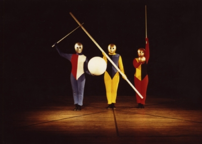Gestentanz performance. Three performers stand in line in front of the viewer. They are wearing multicolored jumpsuits. All of them are wearing golden masks that cover their entire head. The one on the left holds a wand in his right hand and a white sphere. The performer in the middle has a pole diagonally across the front of his body. The one on the right holds a small golden sphere in his right hand and a wand in his left hand. The wand is pointing up.
