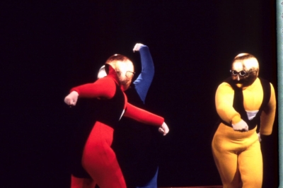 Gestentanz performance. Three performers dance on stage with their arms extended to the sides. They wear red, blue, and yellow jumpsuits from left to right. Their costumes also include golden masks that cover their heads. The masks have round eyeglasses and mustaches.
