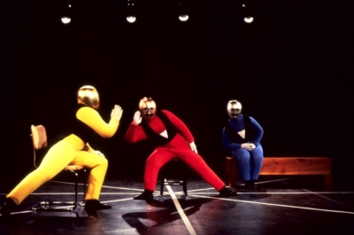 Gestentanz performance. Three performers are sitting on the stage, facing the viewer. They wear yellow, red, and blue jumpsuits from left to right. All the performers wear a golden metallic mask covering their heads and a black tailcoat. The masks have round eyeglasses and mustaches. The performer on the left is leaning towards the performer in the middle, trying to tell him something. The performer in the middle leans towards the performer on the left with his hand close to his ear.