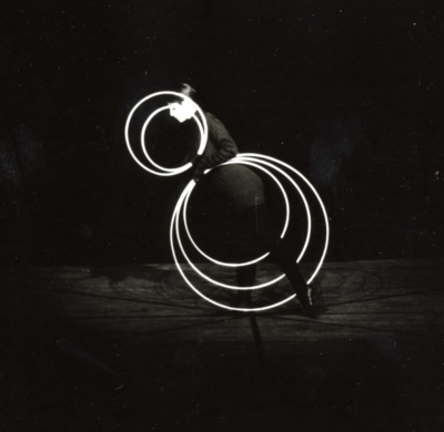 Reinfentanz performance. A black-and-white image of a performer facing the left of the image shows her standing on one foot, with her left foot stretched towards the back. She is holding five white rings in her hands. Two of the rings make concentric circles near her head, and the other three rings form concentric circles below her hips.