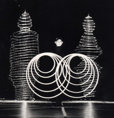 Reinfentanz performance. This is a black-and-white image of a performer standing and facing the viewer. She holds five rings in each hand, making ten interlocking concentric circles before her.