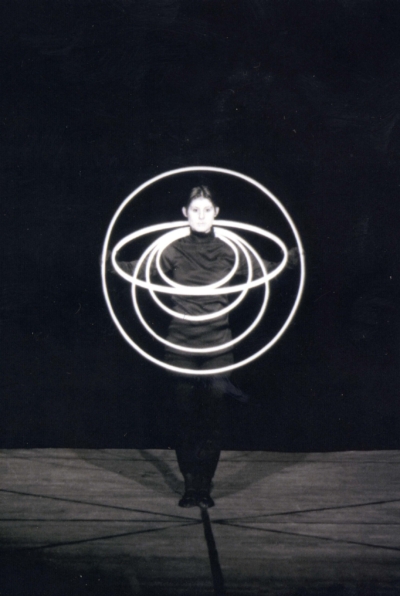Reifentanz performance. This is a black-and-white image of a performer dressed in black standing at the center of the stage. She has five white rings around her neck. She holds one of them to make a circle that frames her head and torso.