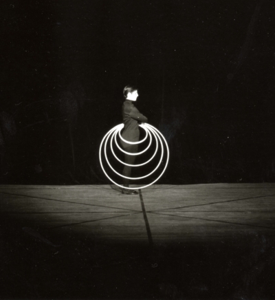 Reifentanz performance. This is a black-and-white image of a performer standing in profile, looking to the right of the image. She is cross-armed and holding five hoops at her hip. The hoops have increasing diameters, with the largest one making a circle from her hip to her feet.