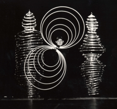 Reinfentanz performance. This is a black-and-white image of a performer. She is standing and facing the viewer. She is holding ten rings close to her face with her hands. The first five rings make concentric circles above her head, framing her face, and the other five rings make concentric circles below her head, slightly to her right. Two anthropomorphic wire shapes in the background are part of the set. These wire shapes are taller than the performer.