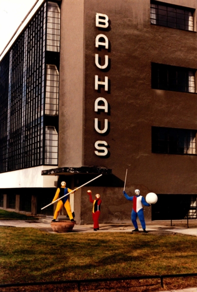 Three performers in colorful costumes in front of a building. The word Bauhaus is written vertically in front of the building.