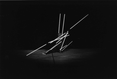 Staebetanz performance. Black-and-white image of a hooded figure crouching away from the viewer. Arms are extended to each side of the dancer. The left leg is extended to the back, and the right is bent at the knee. Twelve white poles of various lengths are attached to the dancer's body, forming straight lines that extend from the limbs and torso. The poles create a strong diagonal that follows the extended arms.