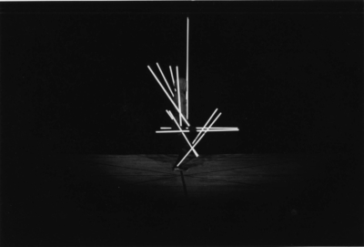 Staebetanz performance. Black and white image of a hooded figure standing in profile, facing towards the right of the image. Twelve white poles of various lengths are attached to the dancer's body, forming straight lines that extend from the limbs and torso. Some poles are vertical, others horizontal, and others are diagonal, following lines from the hip to the knees or upward along the shins.