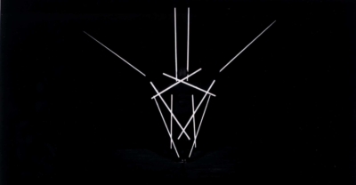Staebetanz performance. This is a black-and-white image of a performer holding twelve poles. The image is underexposed, showing only the bright poles against the dark background. The dancer's poles make a symmetrical pattern radiating from center of the image.