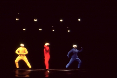 Baukastenspiel performance. Three dancers are on the stage. All dancers are wearing golden masks that cover their entire heads. The dancer on the left is wearing a yellow jumpsuit. He's leaning to his right, with his right knee slightly bent and his left leg straight. The dancer in the middle is wearing a red jumpsuit. He's standing in profile. The dancer on the right is leaning forward, facing toward the right of the image. His left knee is slightly bent.